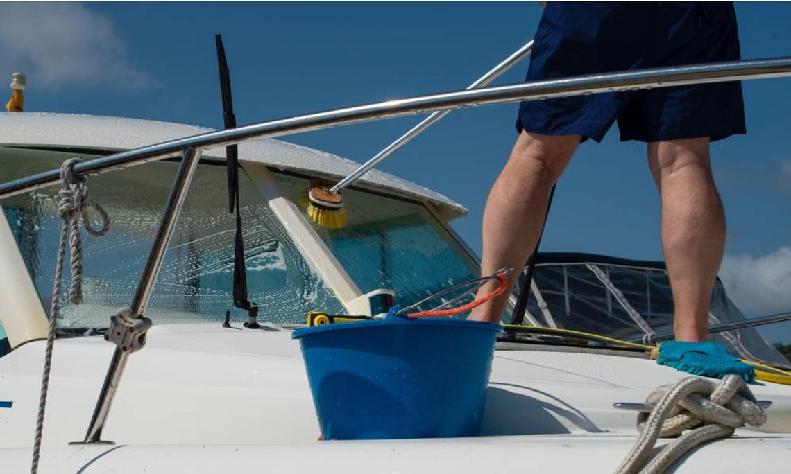 A man scrubbing a white boat using a brush while a pressure water system sprays water with a clear sky in the background.