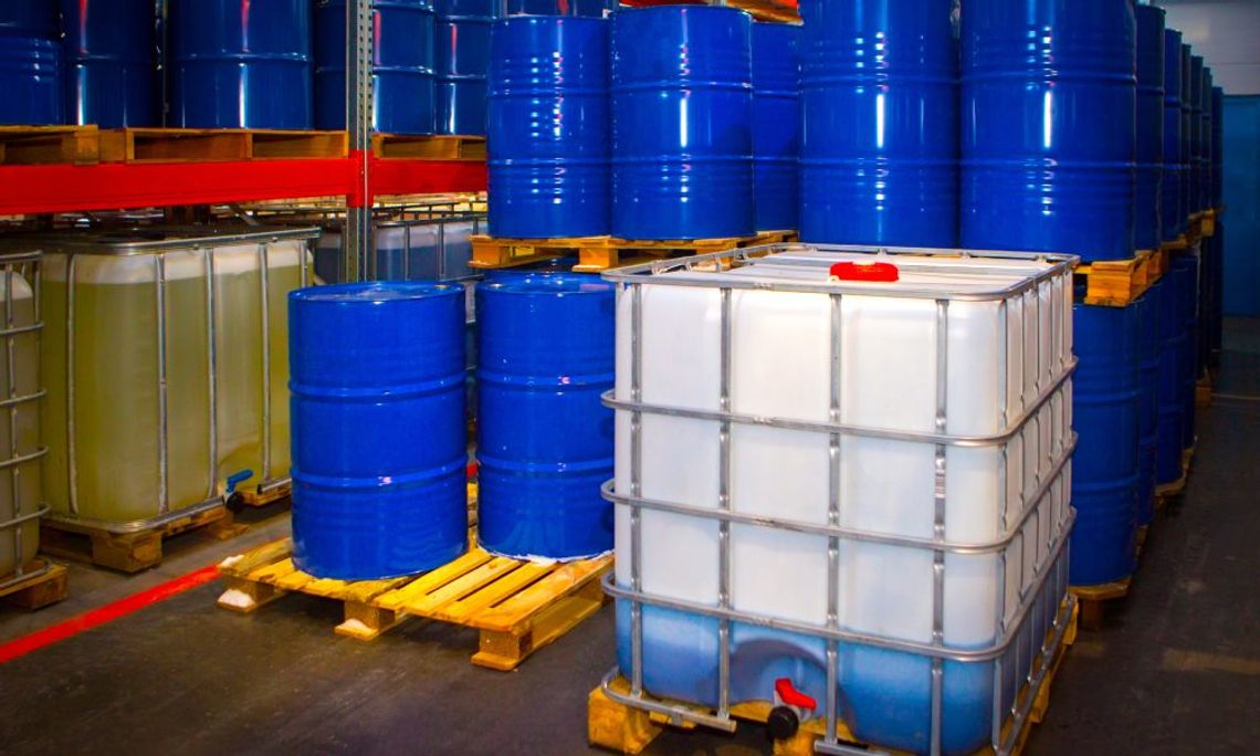 Several blue metal barrels are organized and stacked in an industrial warehouse. A clear, plastic container sits near them.