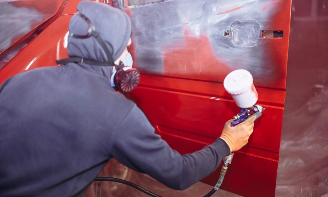 A person wearing a gray hoodie and a respirator mask uses a spray paint gun to paint a damaged car red.