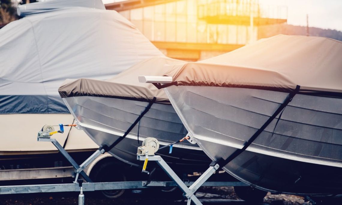 A close-up of a row of boats, each covered with a fabric boat cover. They are lined up in front of a storage building.