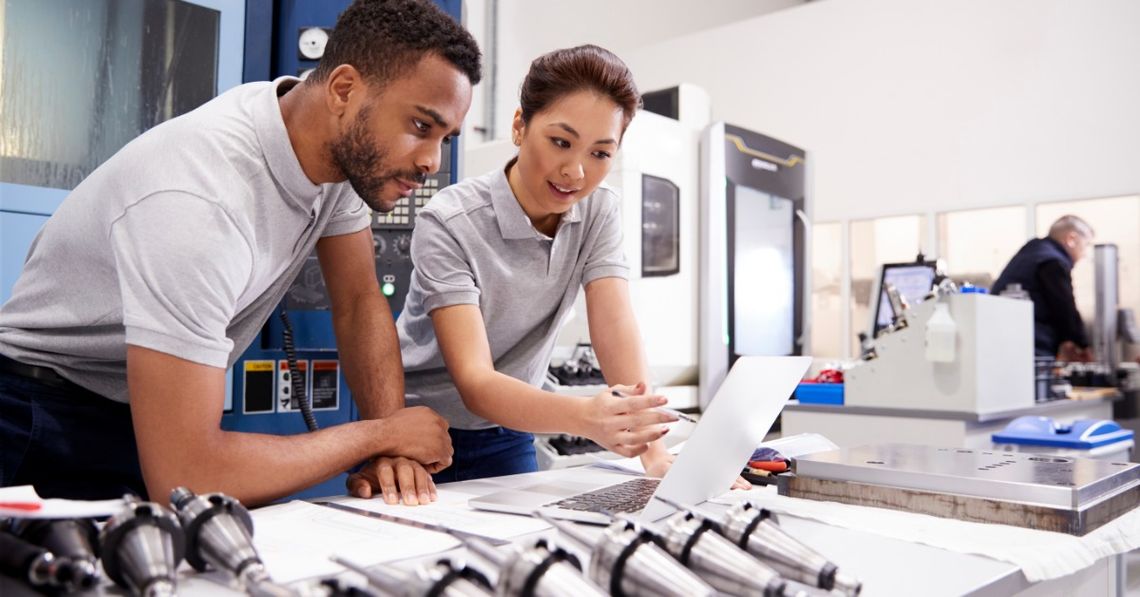 Tips for Improving Process Validation in Product Development Two engineers wearing gray shirts look at a laptop on a large table in a bright manufacturing facility.