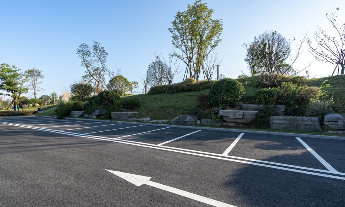 A black asphalt parking lot on a sunny day. The white line striping is fresh and crisp, and there are trees nearby.