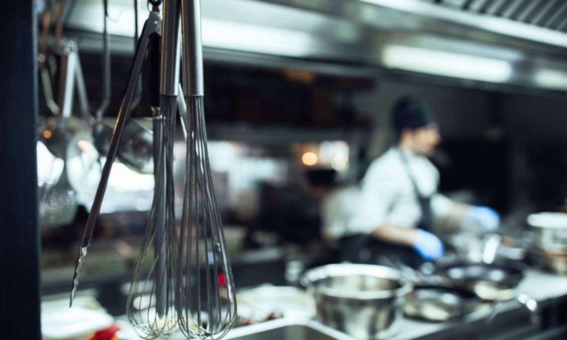 Metal utensils hanging in a commercial kitchen behind a restaurant. A chef is cooking on a stovetop nearby.