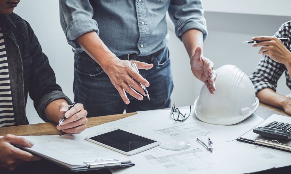 Three individuals at a wooden table with building models, tools, and blueprints spread out in front of them.