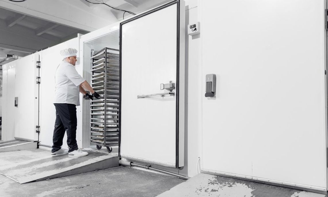 A man wearing a white uniform and a chef's hat, pushing a metal cart with stackable trays inside an industrial freezer.