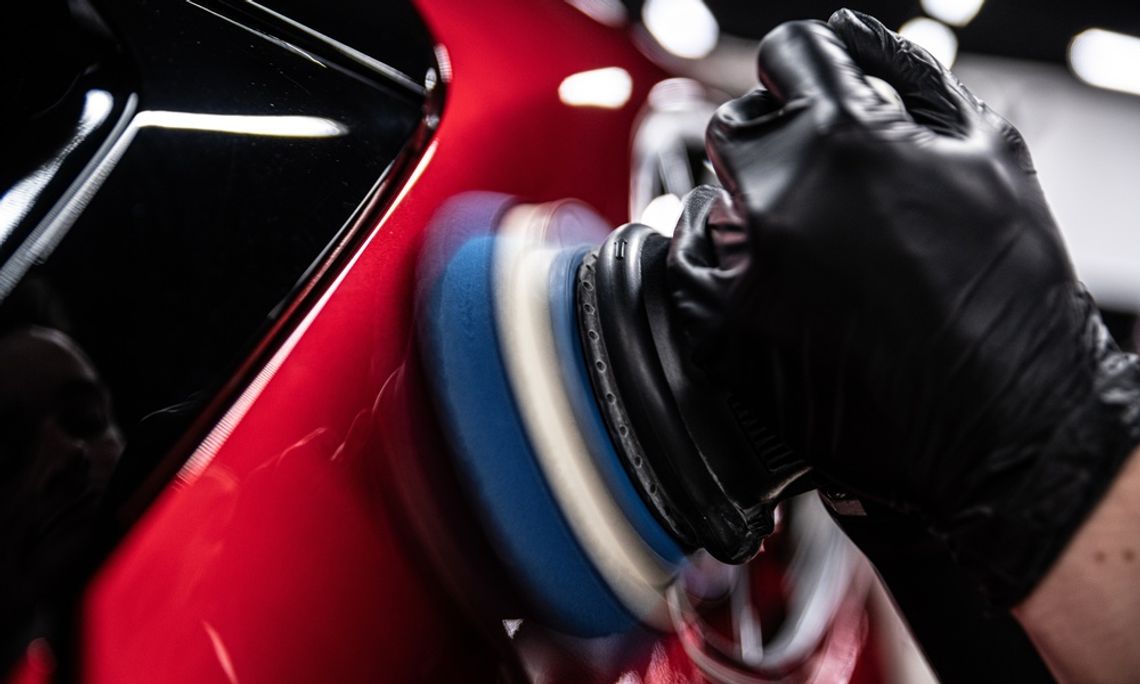 A close-up of a professional using an electric machine for paint correction in a red car detailing studio.