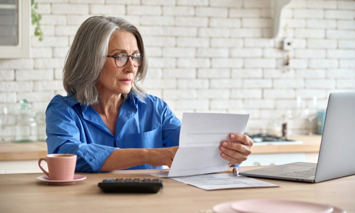 An older woman sits at the kitchen counter looking at a piece of paper. A laptop, a mug, and a calculator are on the counter.