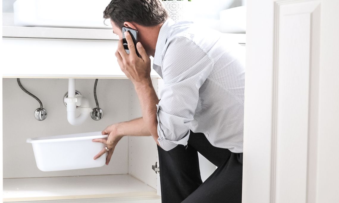 A homeowner kneeling under his bathroom sink, holding a bucket to catch water. He's talking on the phone with the other hand.