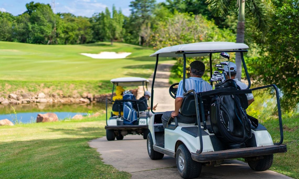 A group of four people in two separate golf carts is riding through a golf course cart path near a faux lake.