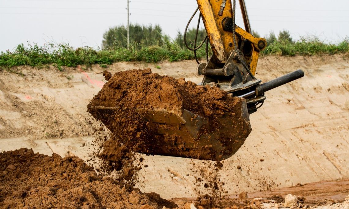 A yellow excavator bucket scoops clay-rich soil at a construction site with dirt mounds and vegetation in the background.