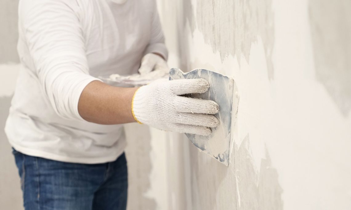 Person wearing gloves applies joint compound to a drywall surface with a taping knife while smoothing wall repairs indoors.