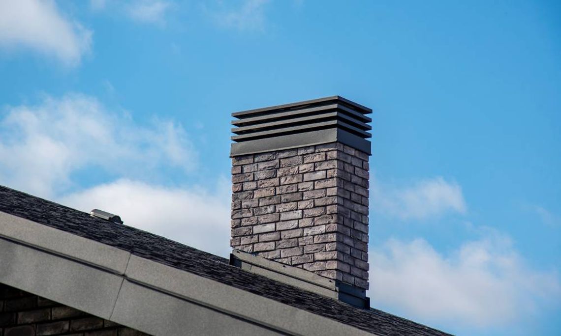 Understanding the Benefits of Custom Chimney Caps A brick chimney with a black custom cap above a shingled roof under a bright blue sky with soft clouds.