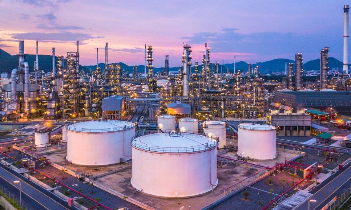 An aerial view of a large oil and gas refinery at twilight. The refinery has several chemical tanks and vertical pipes.