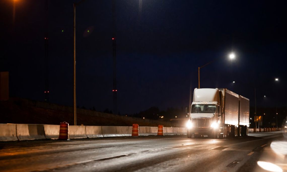 A white semi-truck with its headlights on driving next to road barricades and orange and white traffic barrels at night.