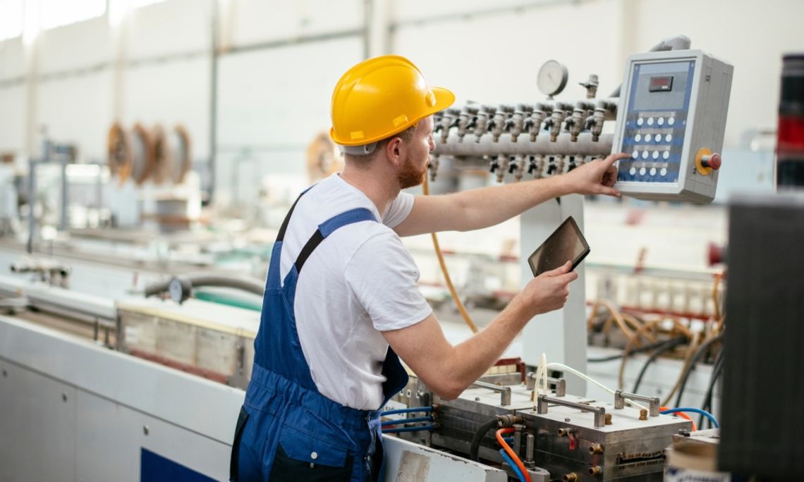 A male factory worker operating machinery using a tablet and a control panel with a digital display.