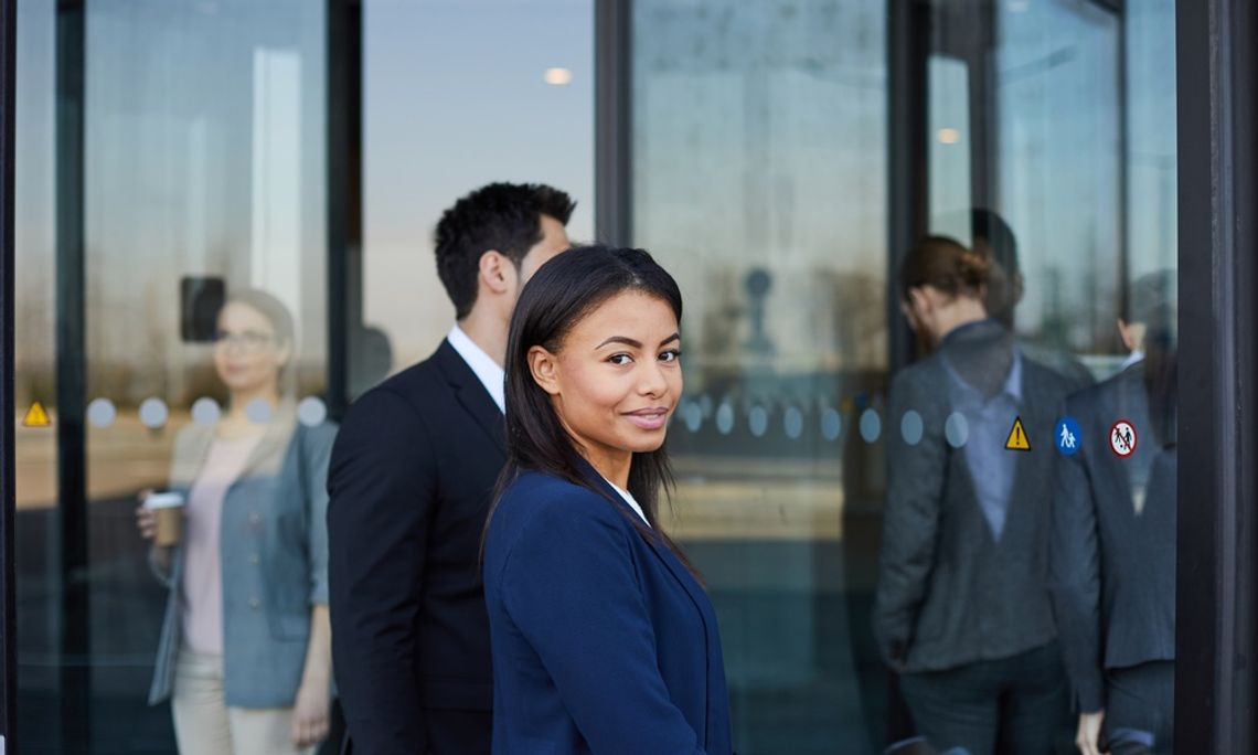 A woman dressed in business professional attire looks to the side while walking through an automatic door.