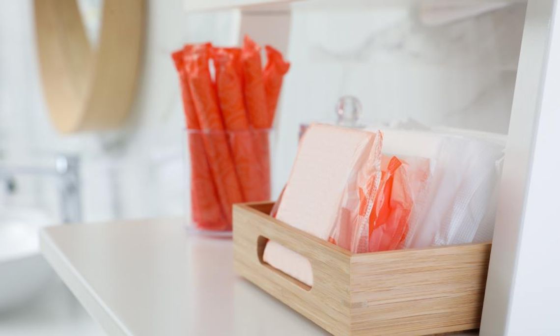 A small wooden crate contains menstrual pads of various sizes. A glass jar contains tampons in orange wrappers.