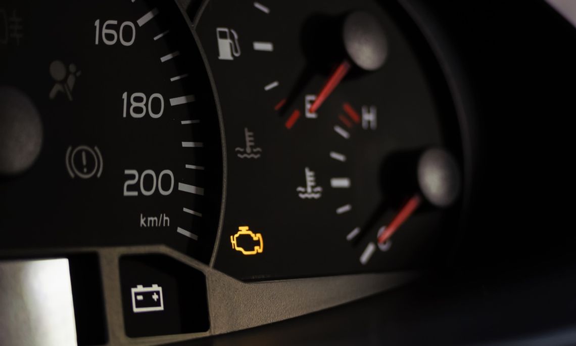A close-up focos on a vehicle dashboard. A small yellow check engine light displays beside the speedometer.
