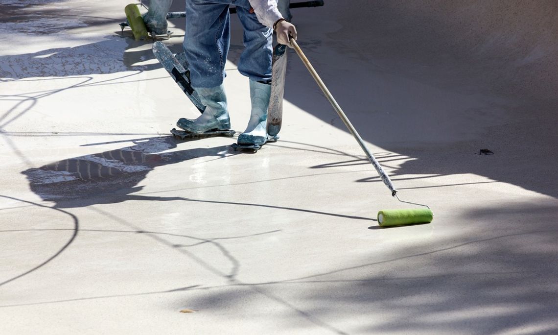 A worker in rubber boots uses a long-handled roller to apply coating to an empty concrete swimming pool surface.