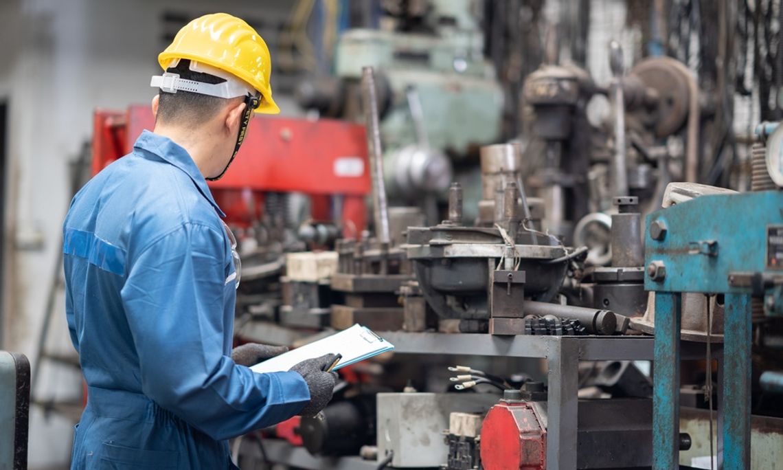 A person wearing a yellow hard hat is holding a small clipboard and inspecting metal machine components.