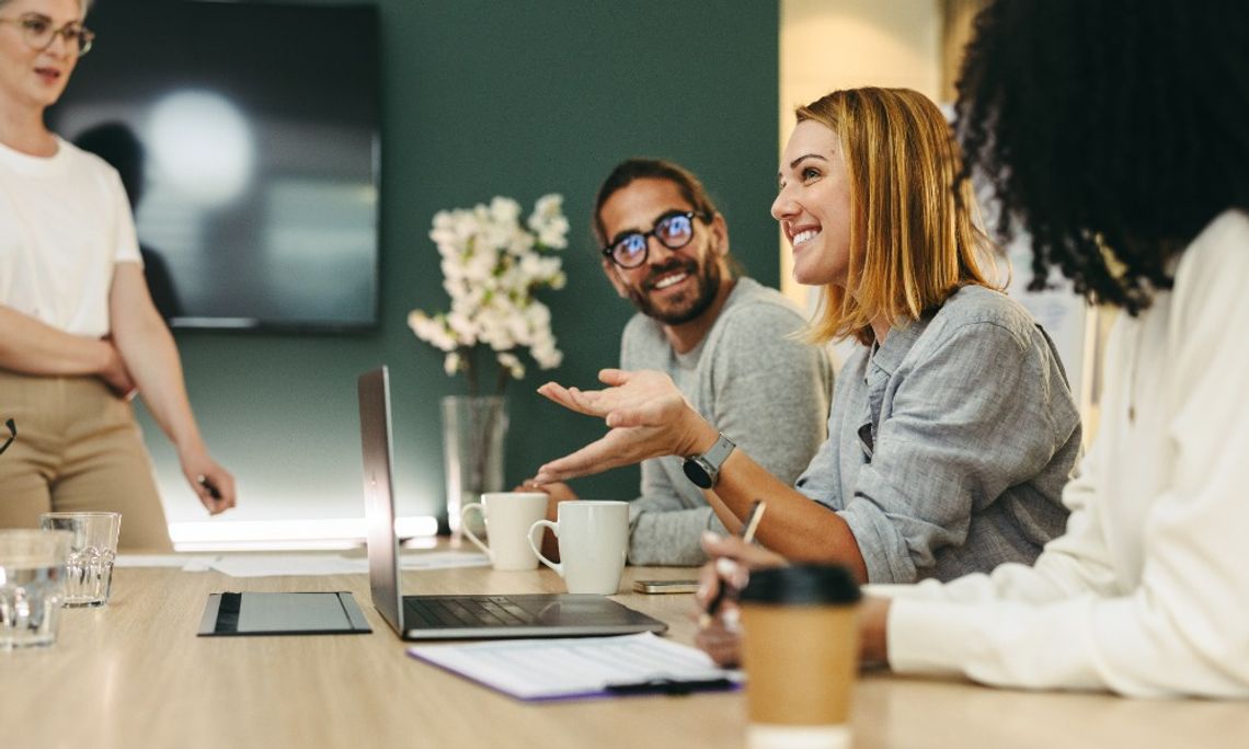Coworkers gather around a conference table with laptops, coffee cups, and papers. One person gestures while speaking.