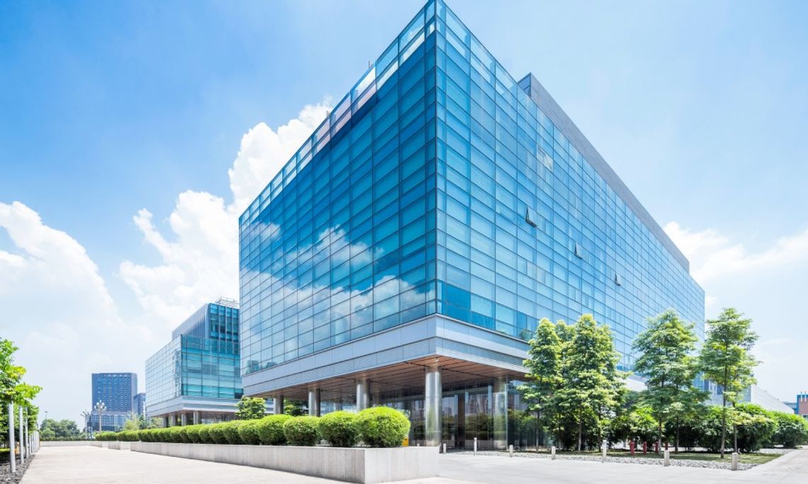 A glass commercial building sits next to a glass commercial building. There are mature shrubs and trees on the property.