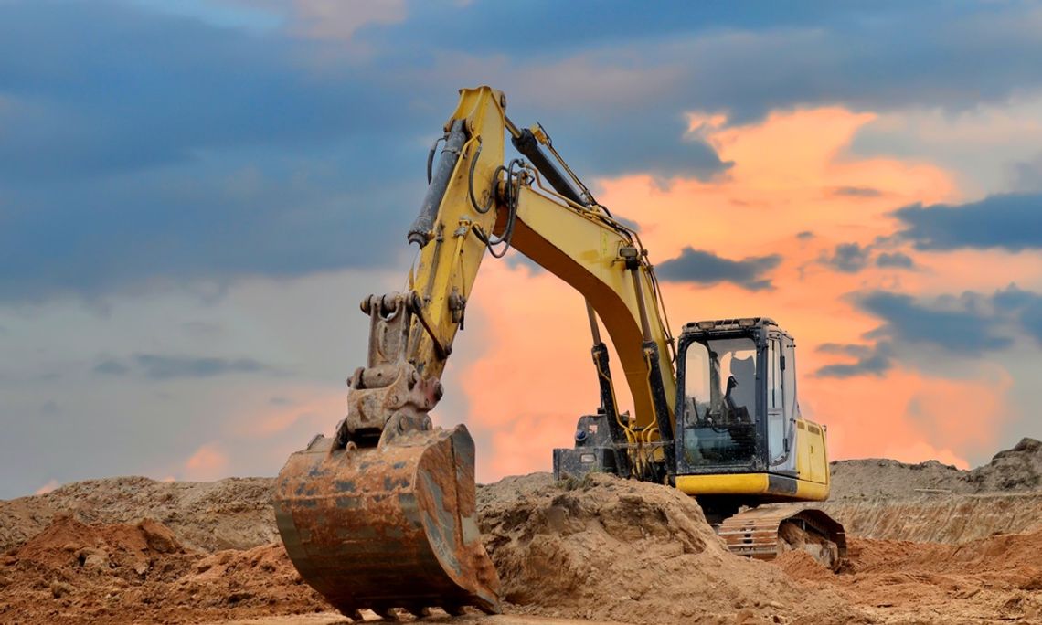 What Contractors Check Before Drilling Starts A yellow excavator digging into large mounds of brown soil beneath a cloudy sky with orange light near the horizon.