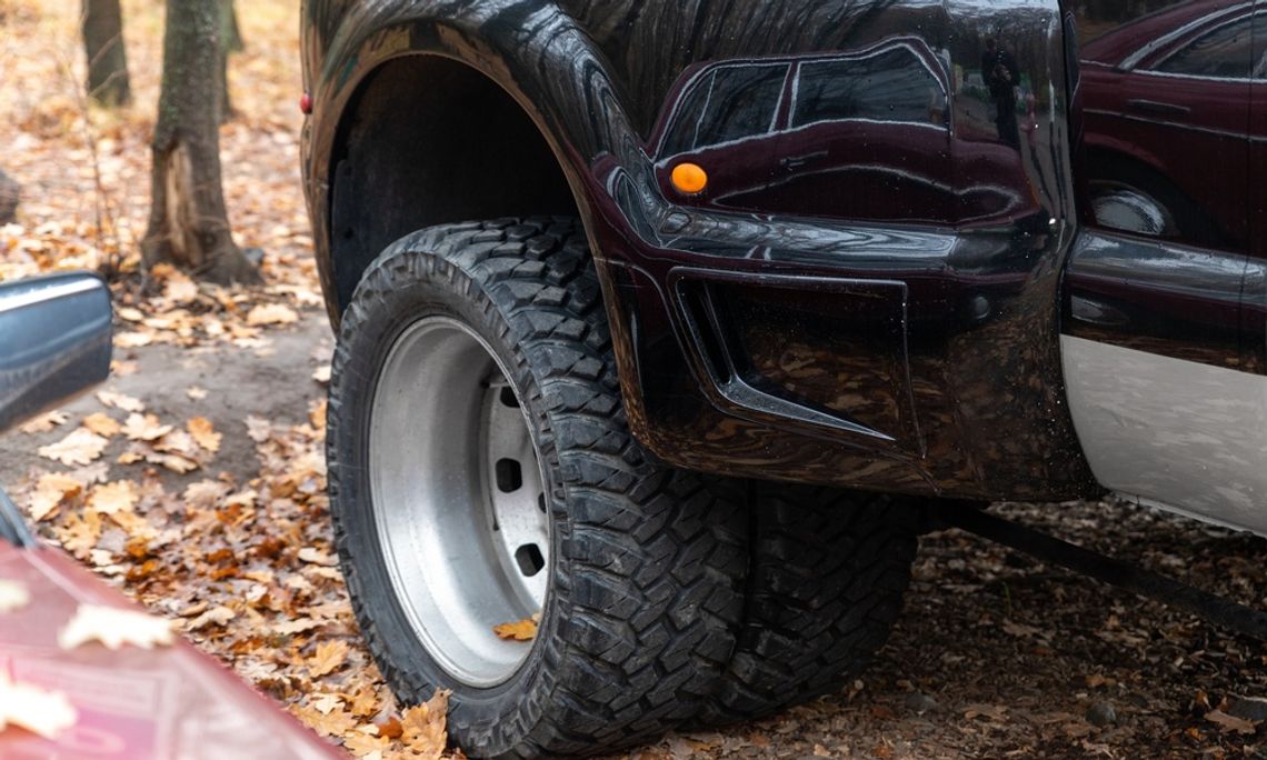 A close-up of the back tires of a pickup truck with dually wheels parked in a forest over dry leaves next to trees.
