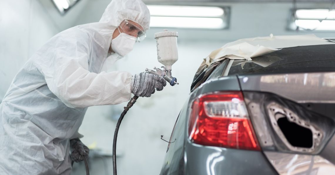 What Happens Before a Car Gets a Fresh Repaint A person in protective gear spraying a gray car body with a paint gun inside a bright automotive paint booth.