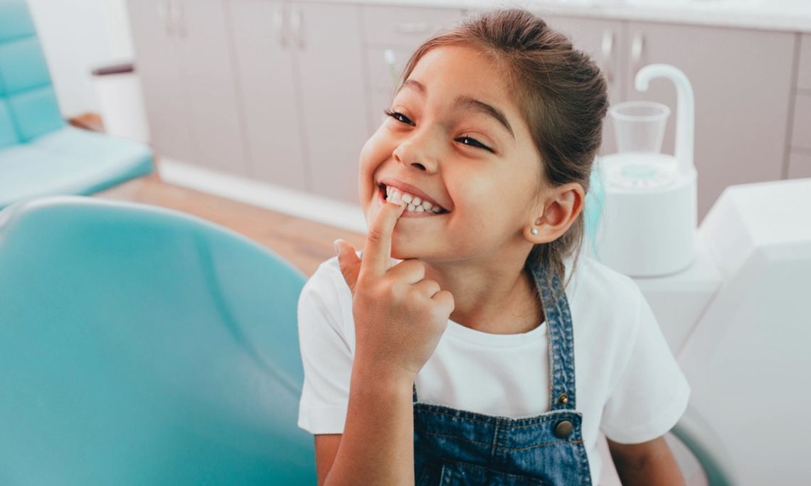 A young girl in a white shirt and overalls smiles while pointing to her teeth in a dentist's chair in an office.