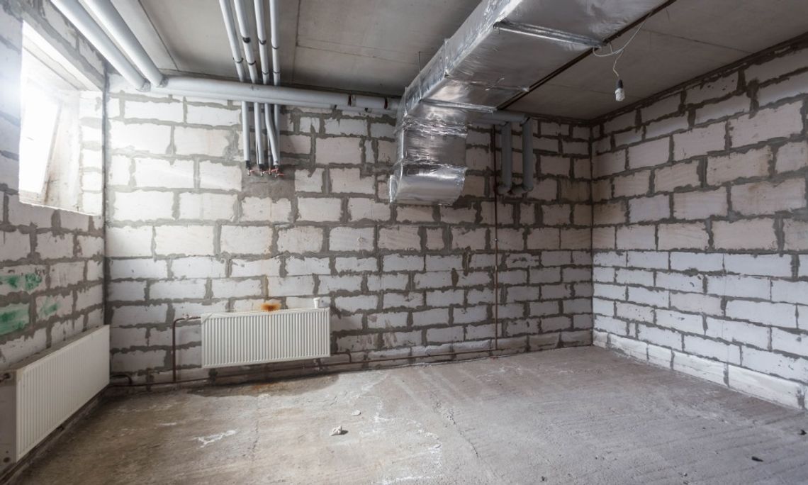What To Check Before Finishing an Older Basement An unfinished basement with exposed concrete block walls, overhead pipes, HVAC ductwork, radiators, and a bare concrete floor.