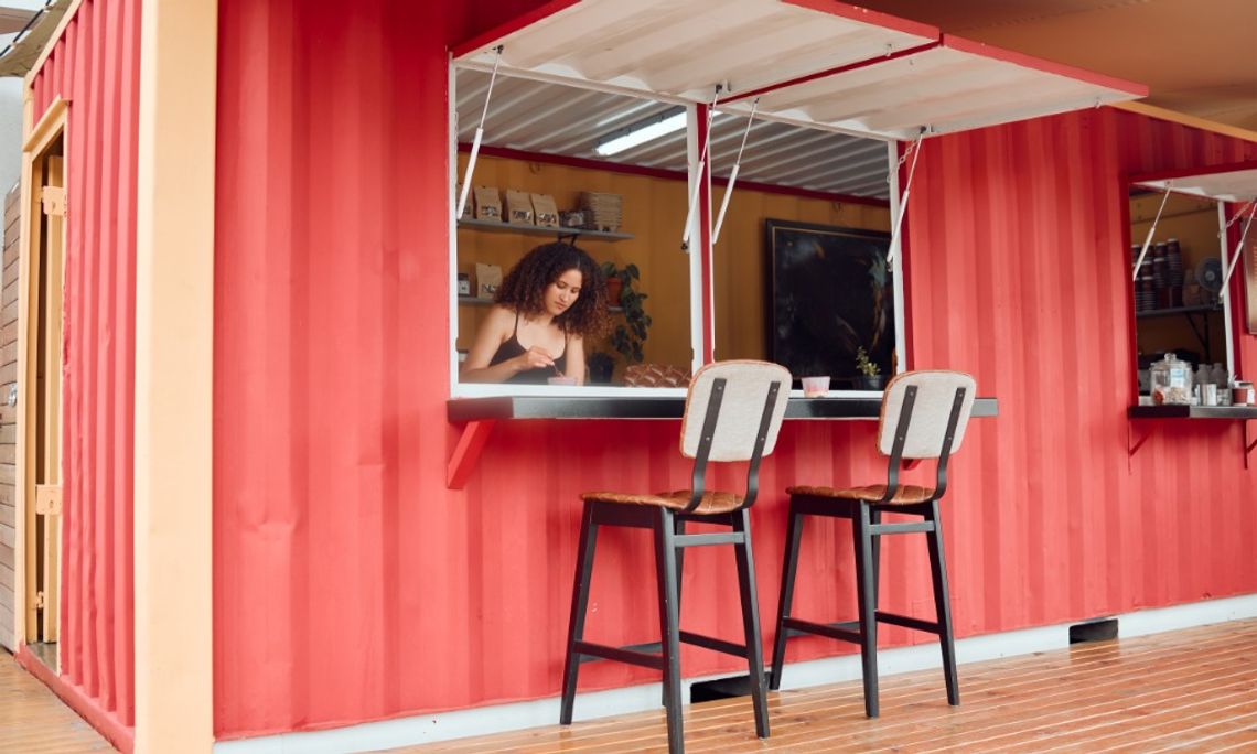 Woman working inside a red shipping container restaurant with an open service window and two stools outside.
