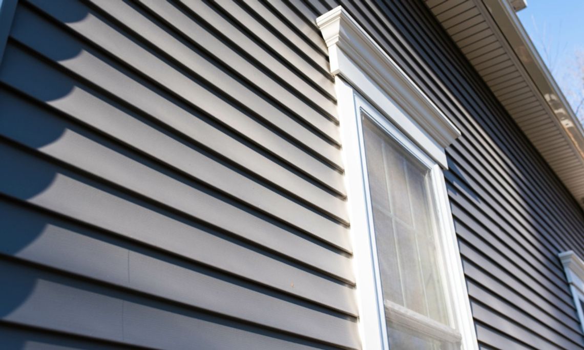 Dark gray horizontal siding covers a home exterior beside a tall window with white trim under a roof overhang.
