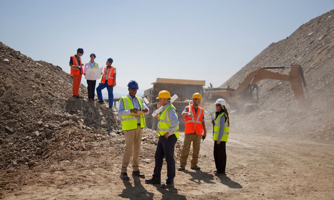 What To Consider When Updating Your Crew’s Workwear Several construction workers in hard hats and safety vests reviewing plans at a rocky excavation site with heavy machinery.