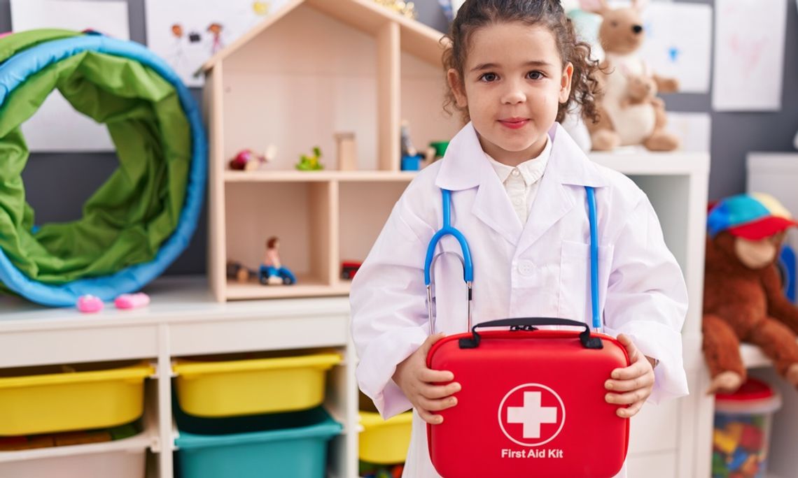 A little girl dressed up as a doctor and holding a red first aid kit in front of her as she stands in a classroom.
