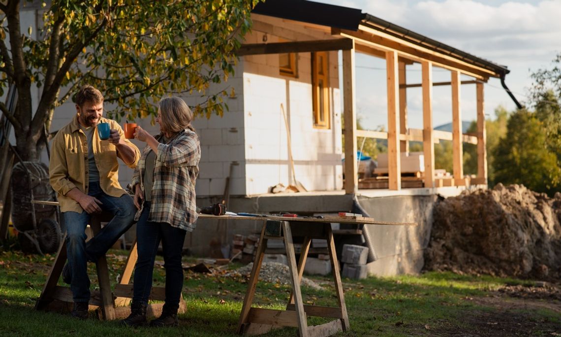 What To Expect During an Exterior Home Remodeling Project People holding mugs near a work table and trees. There is an unfinished house with wood posts behind the couple.