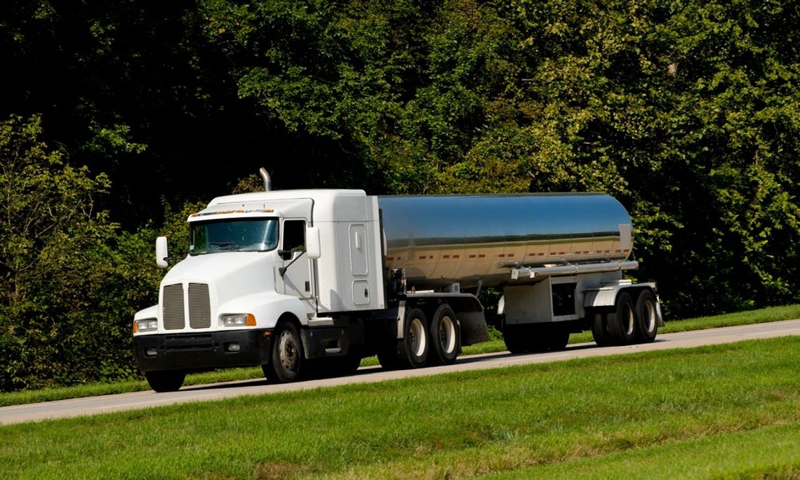 A tanker trailer with a white cab and silver fuel tank drives along a road between a treeline and grassy divider.
