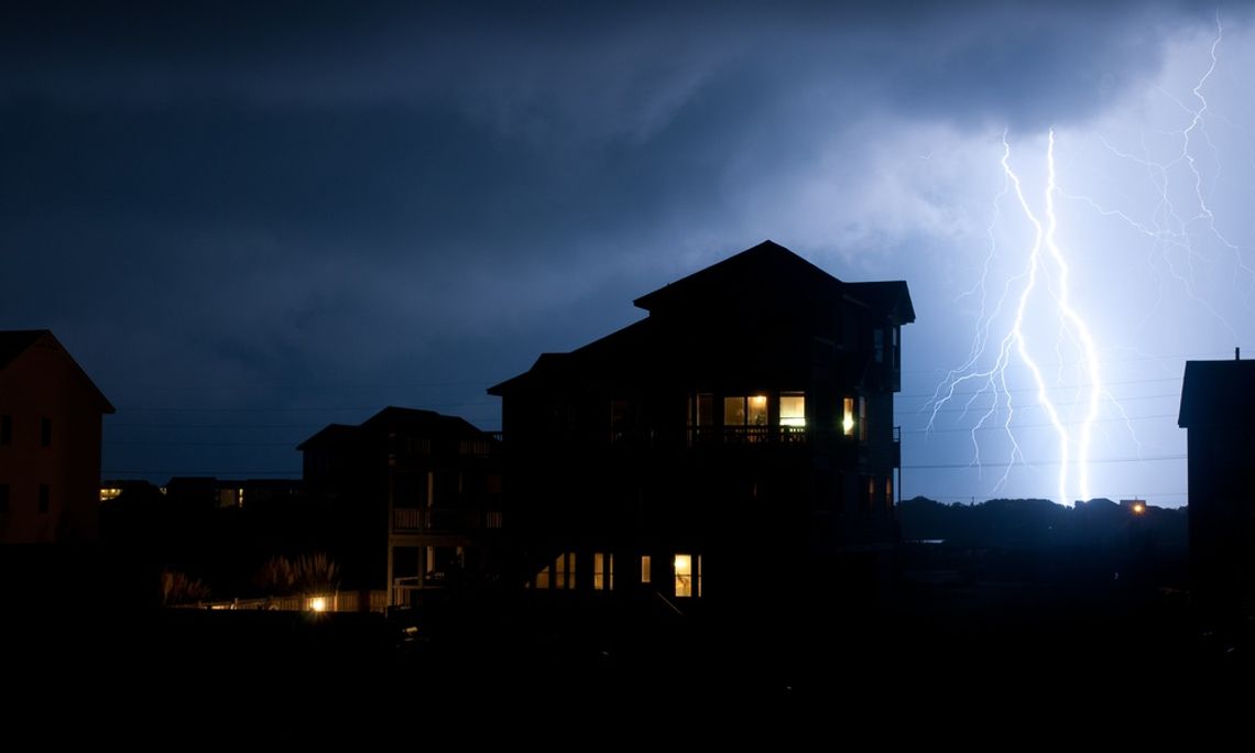 A silhouette of a house on a stormy night. There are two large lightning bolts travelling down to the ground behind it.