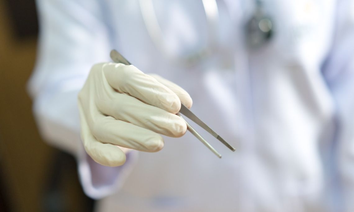 Gloved medical professional holding stainless steel thumb forceps against a blurred white coat background.