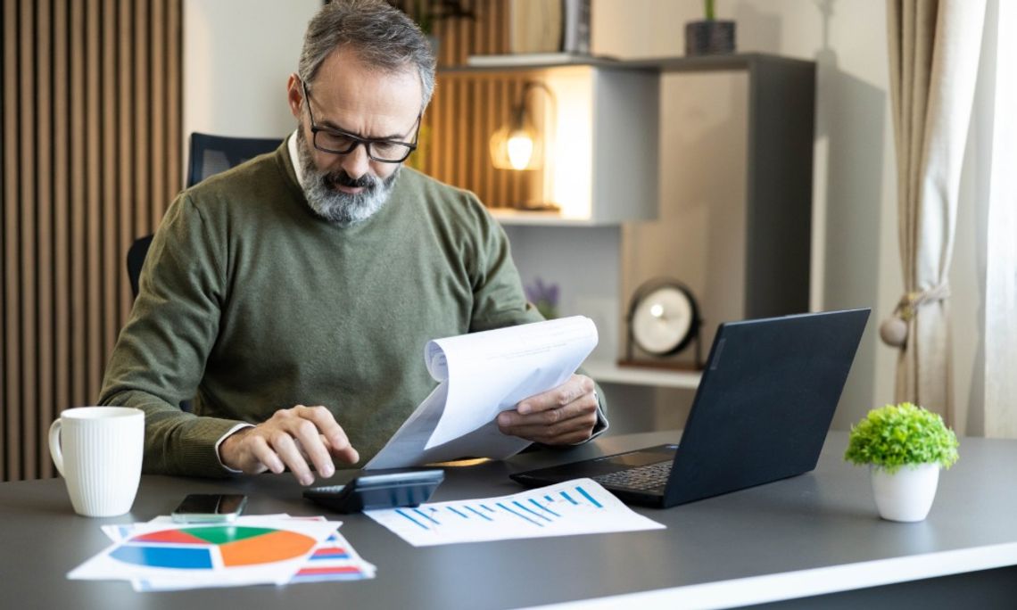 A business owner reviews financial paperwork with a calculator and laptop while charts sit across his office desk.