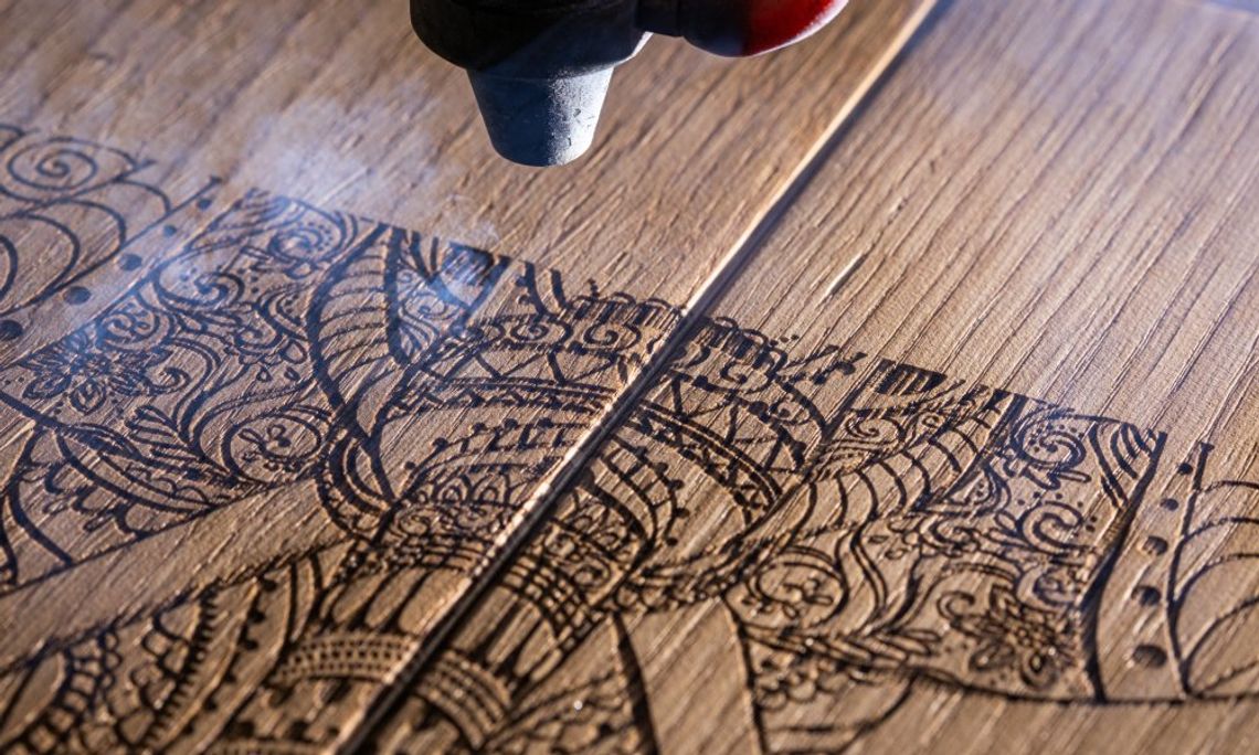 A black and red laser engraving machine is engraving an intricate pattern into a piece of light-colored wood.