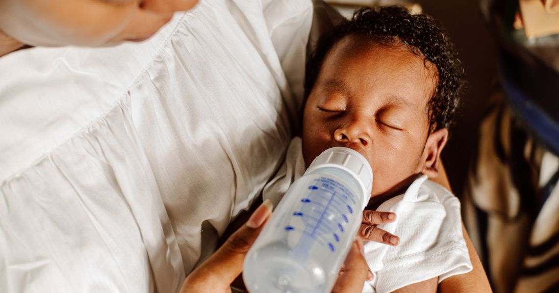 A parent cradles a sleeping baby in her arms. The baby still has an empty milk bottle in their mouth.