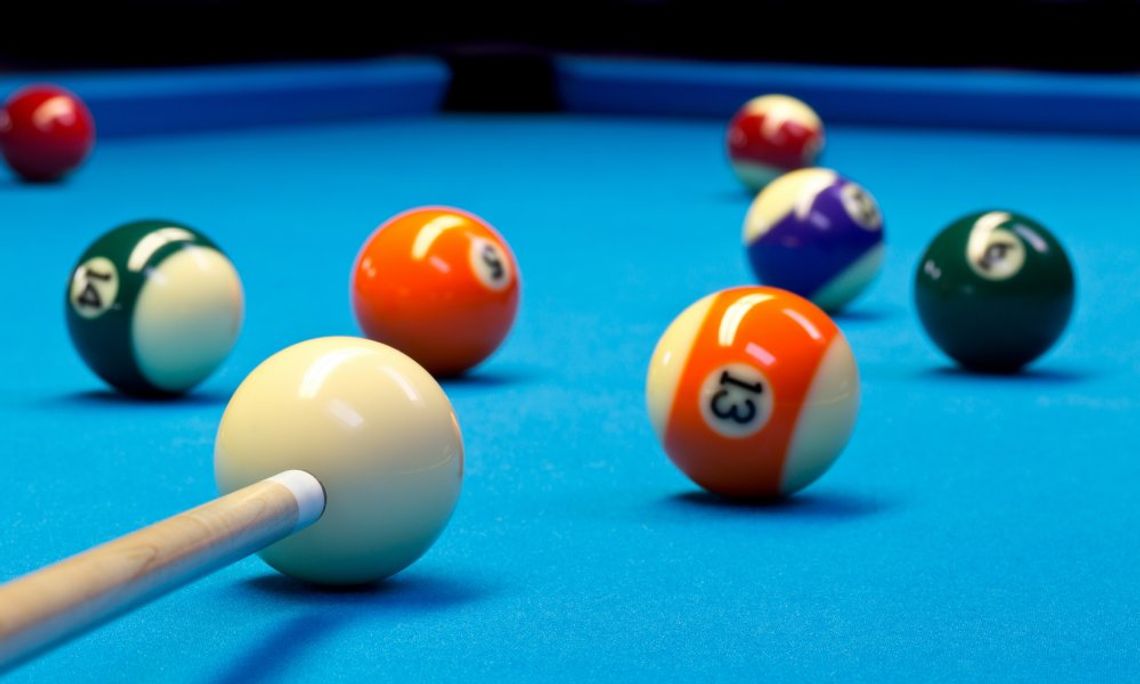 A close-up of a Billiard pool table, and someone is aiming their pool stick towards the orange, striped 13-ball.