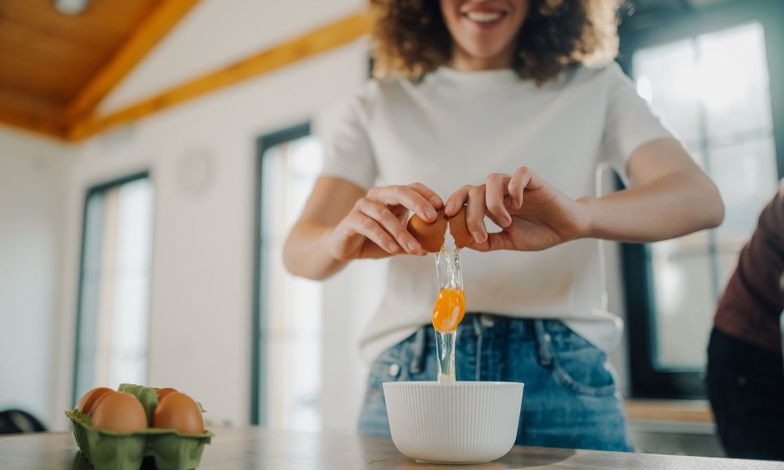 Why Duck Eggs Are Worth Trying in Your Kitchen A woman wearing a white shirt cracks a brown egg into a bowl on a kitchen counter beside an egg carton.