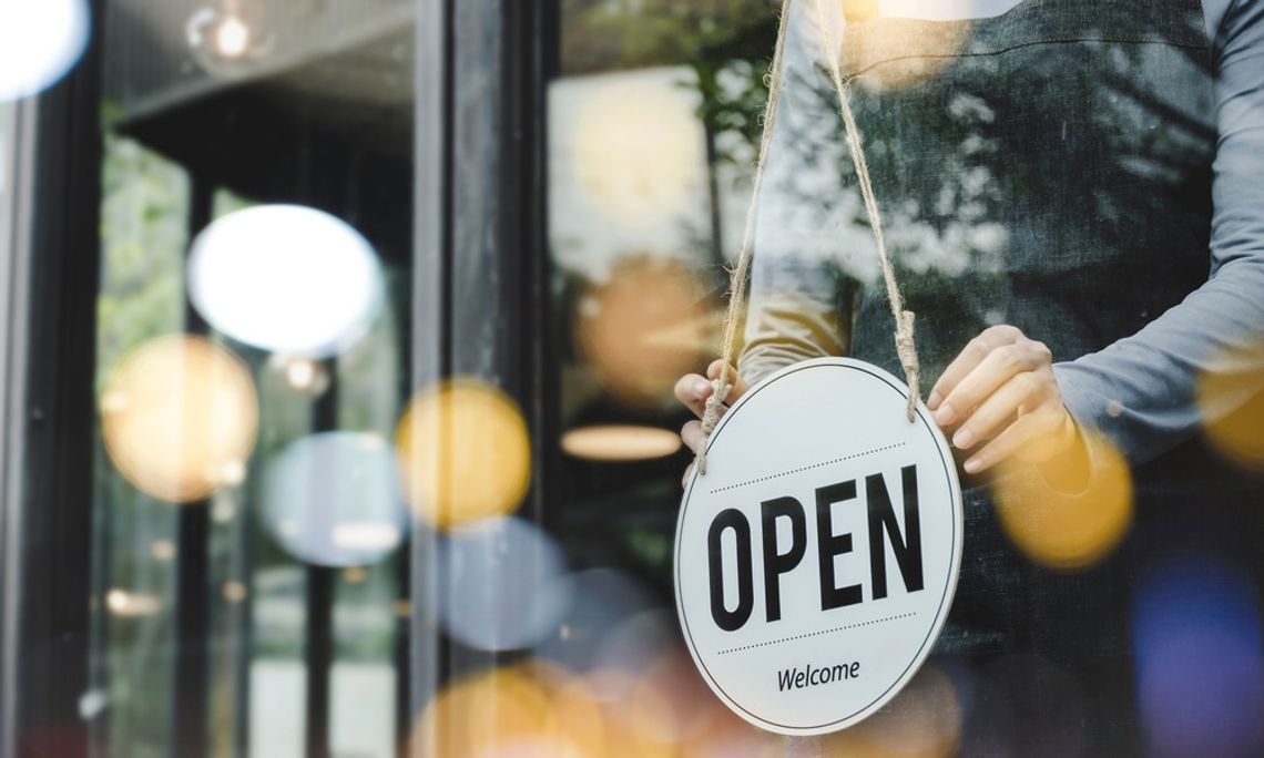 Why Entrance Reliability Matters for High-Traffic Properties A hand turning an open sign on a glass door, with a person in an apron and blurred cafe interior behind it.