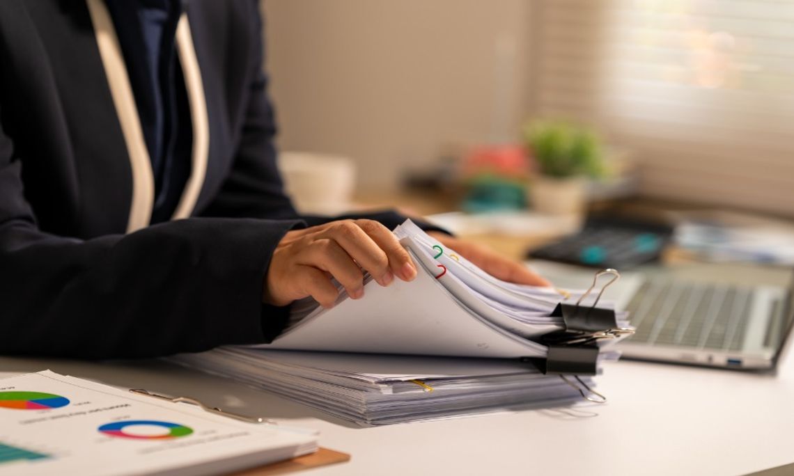 A woman sits at a desk, flipping through papers. There is a laptop blurry in the background.