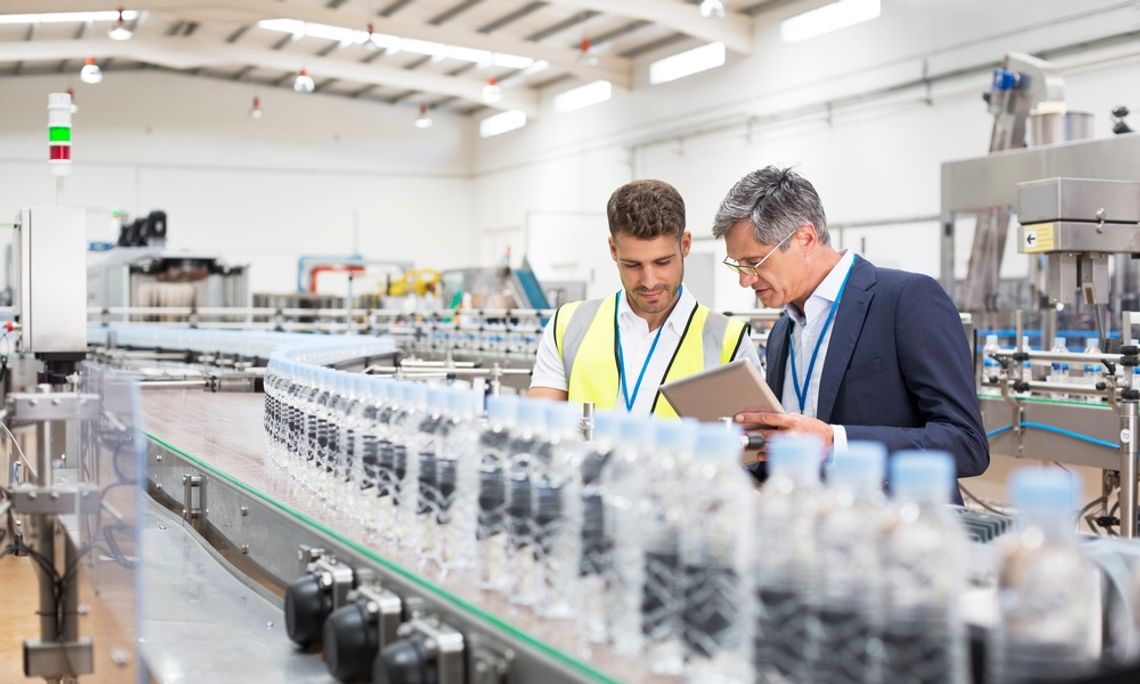 Two men stand behind a manufacturing line with hundreds of plastic water bottles. One person holds a tablet.
