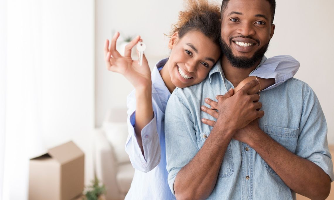 Why New Construction Homes Still Need an Inspection A smiling woman stands behind a smiling man and has one arm draped around his shoulders and the other holding up a key.