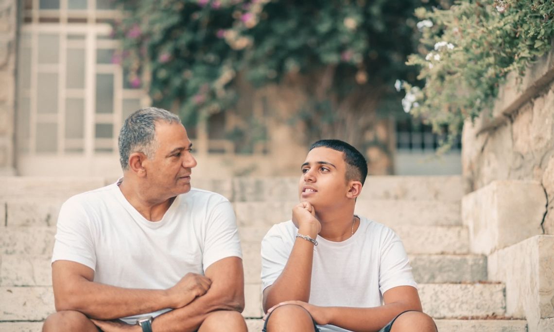 A teenage son looking thoughtfully toward his aging father as they sit on the stone steps of a home and converse.
