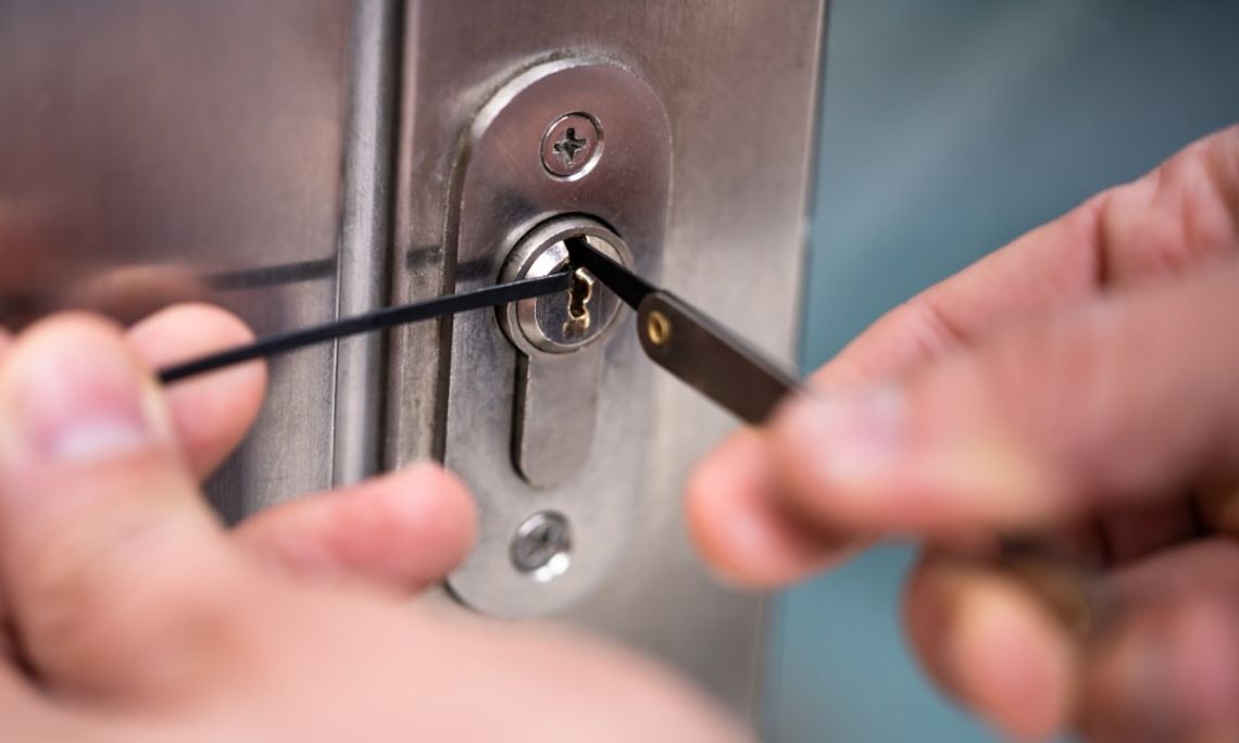 A close-up of two hands holding slender metal tools inside the keyhole of a silver door lock mounted on a metal door.